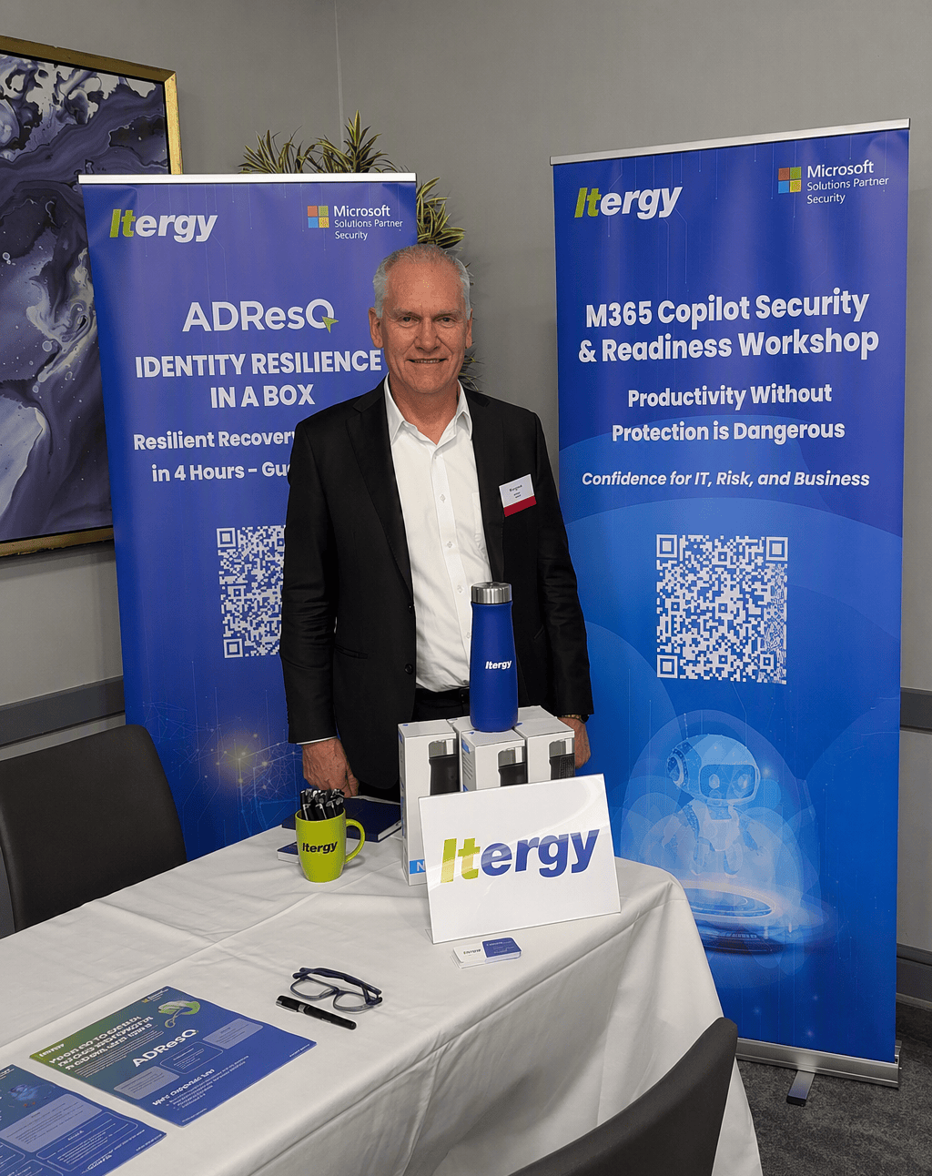 Ralph-event-1 | Itergy Smiling man in a black suit stands at a conference booth between two tall blue banners with Ittergy branding and event titles like 'ADResQ Identity Resilience in a Box' and 'M365 Copilot Security & Readiness Workshop'.